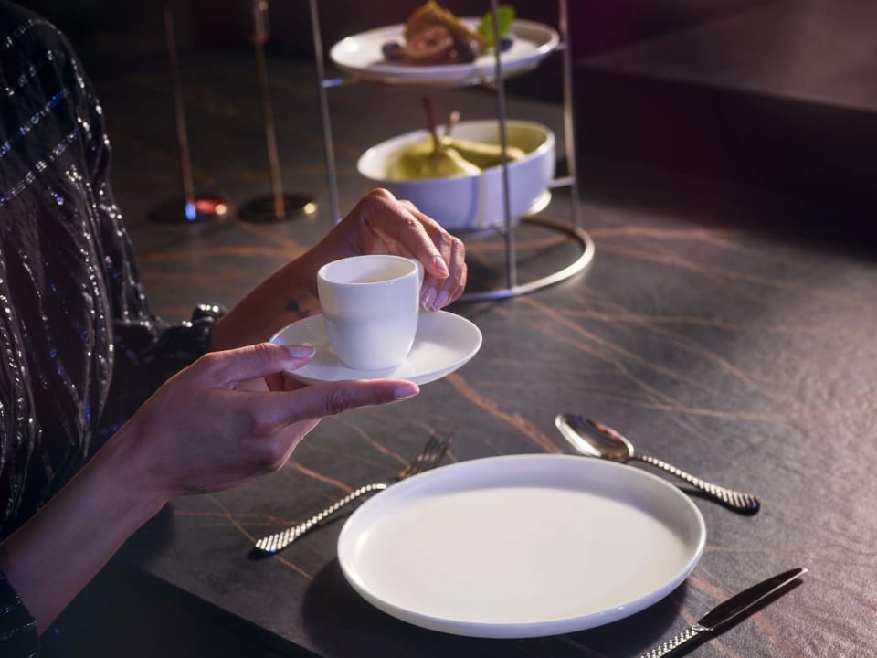 Gedeckter Tisch mit Jade Lift Teller, einer Hand die eine Kaffeeobertasse mit Kaffeunteren h&auml;lt, Etagere im Hintergrund